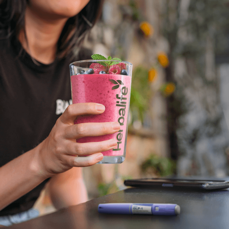 A person holding a Herbalife protein shake glass filled with berry smoothie, showcasing the AuraStylo health drink.